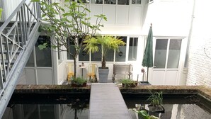 A serene indoor garden with a wooden walkway leading to a small pond, surrounded by potted plants and a seating area with chairs. - Le Comptoir industriel (Lille)