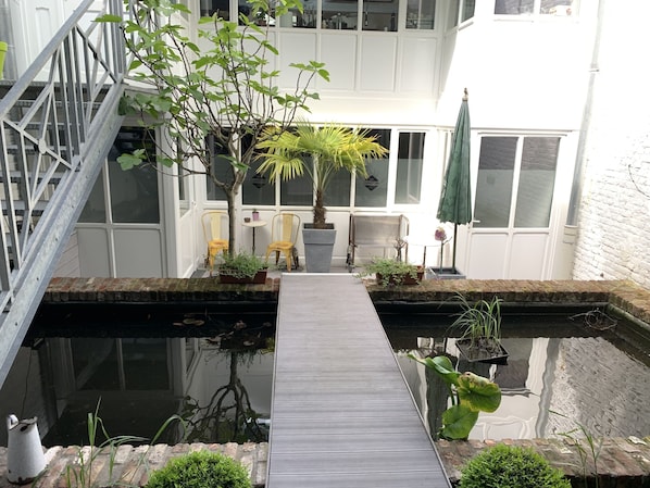 Un jardin intérieur paisible, avec une allée en bois menant à un petit étang, entouré de plantes en pot et agrémenté d’un espace détente avec des chaises.