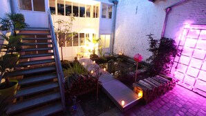 Un espacio exterior bien iluminado con una escalera de madera que lleva a una terraza cubierta, rodeado de plantas en macetas y un pequeño jardín.
