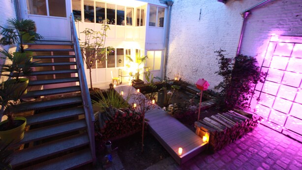 A well-lit outdoor area with a wooden staircase leading to a covered deck, surrounded by potted plants and a small garden. - Le Comptoir industriel (Lille)