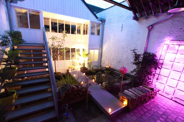 A well-lit outdoor area with a wooden staircase leading to a covered deck, surrounded by potted plants and a small garden. - Le Comptoir industriel (Lille)