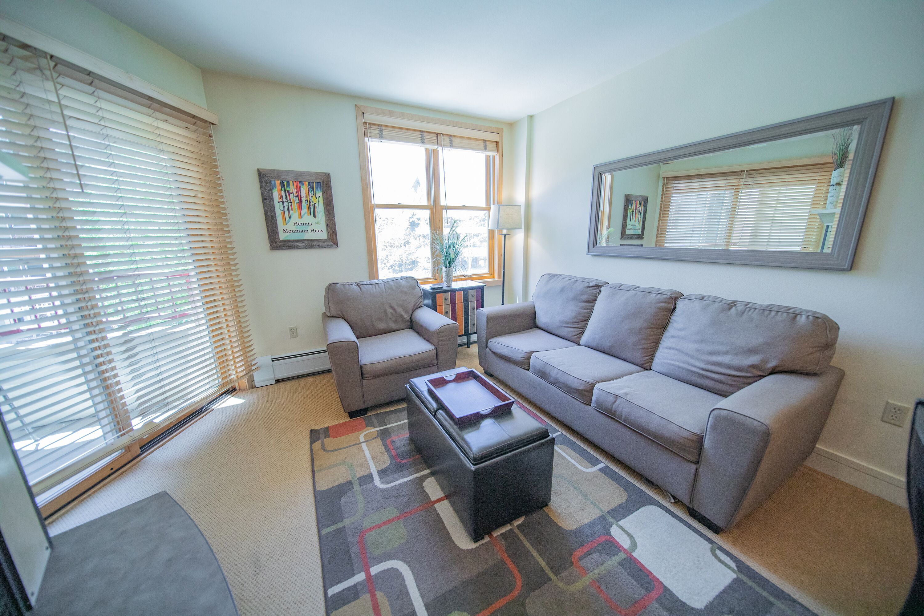 A living room with a sofa, armchair, and ottoman, a patterned rug, and a large window with blinds.