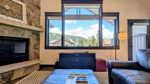 Ein Wohnzimmer mit Kamin aus Stein, einem großen Fenster mit Bergblick, einer schwarzen Ledersofa und einem Couchtisch mit Fernbedienungen.