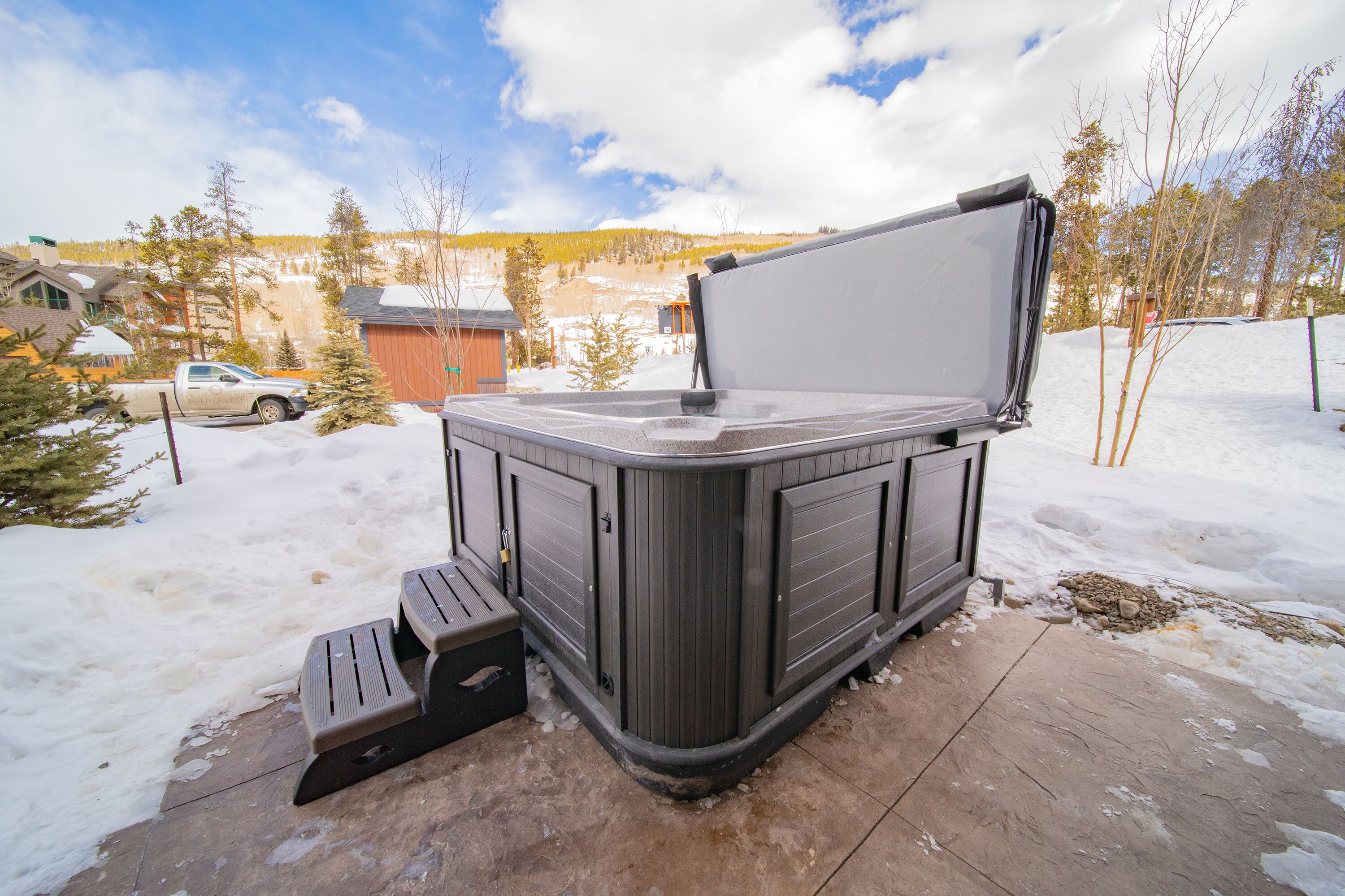 A hot tub with a bench, surrounded by snow, in a residential area with houses and trees.