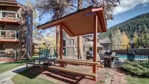 A wooden gazebo with benches, a barbecue grill, and a pool in the background.