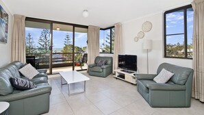 A modern living room with a large sliding glass door leading to a balcony, a flat-screen TV, and a dining area with a white table and chairs. - Sundial 503, 8-10 Hollingworth Street, (Port Macquarie)