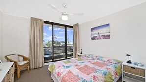 A bedroom with a large bed, a chair, a desk, a ceiling fan, and a window with a view of the beach. - Sundial 503, 8-10 Hollingworth Street, (Port Macquarie)