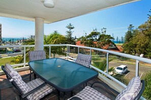 A balcony with a glass table and checkered cushions, overlooking a coastal area with palm trees and a view of the sea. - Mainsail 10, 17 Owen Street, (Port Macquarie)
