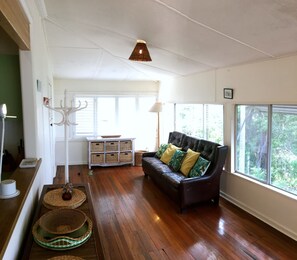 A spacious living room with hardwood flooring, a leather sofa with patterned cushions, a white console table, and a large window offering a view of greenery. - Rosemaree, 24 East Street, (Crescent Head)