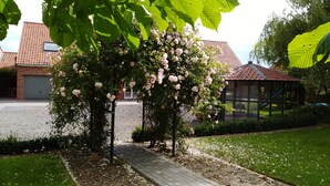 Un sentier de jardin mène à un belvédère orné de roses roses, qui est entouré d’un bâtiment en briques et d’une aire de jeux.