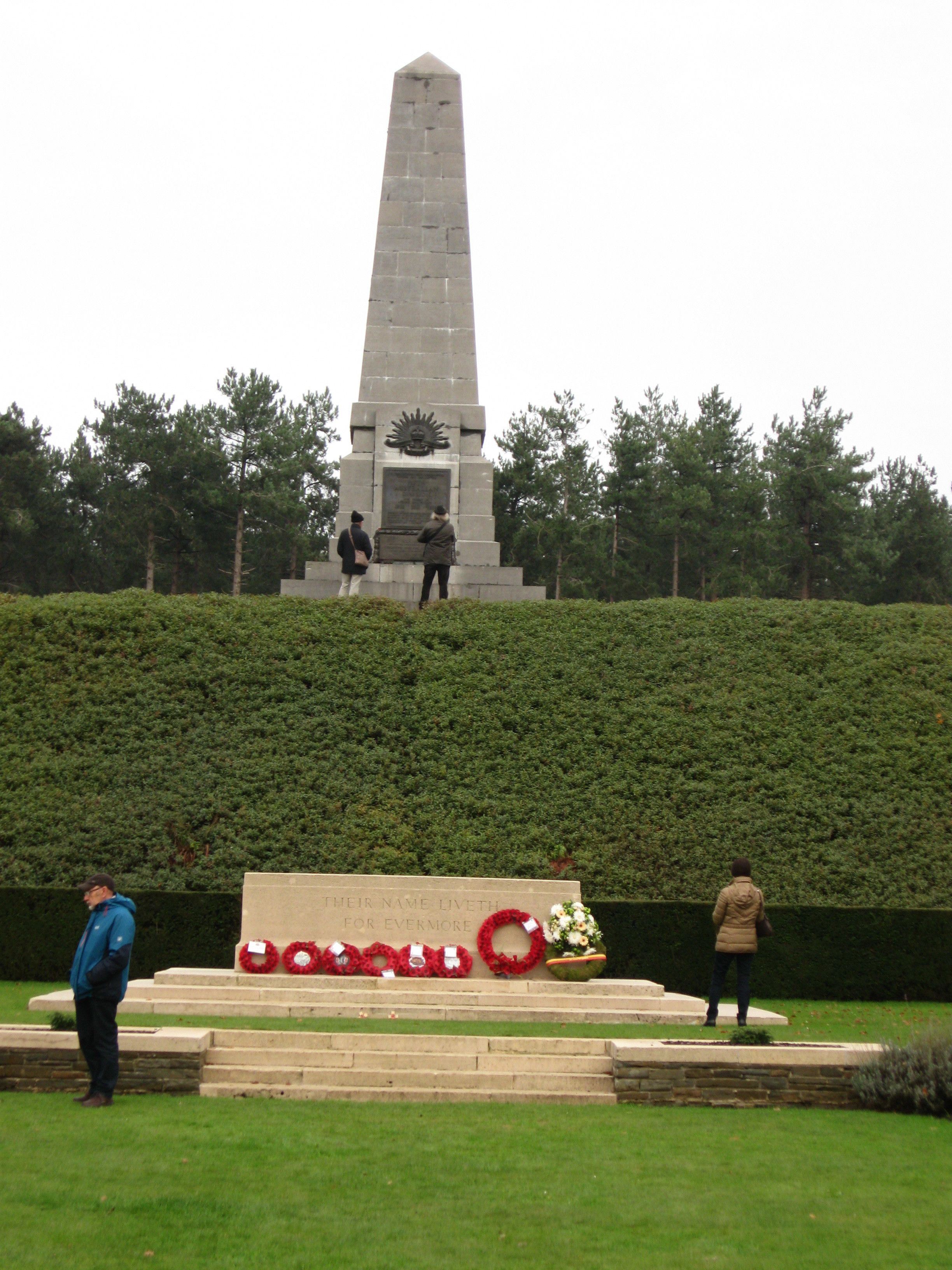 Un monument haut et étroit, surmonté d’une plaque commémorative à sa base, entouré d’une haie bien entretenue et d’une pelouse.