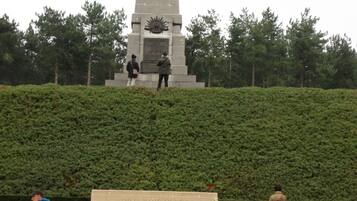 Un monument haut et étroit, surmonté d’une plaque commémorative à sa base, entouré d’une haie bien entretenue et d’une pelouse.