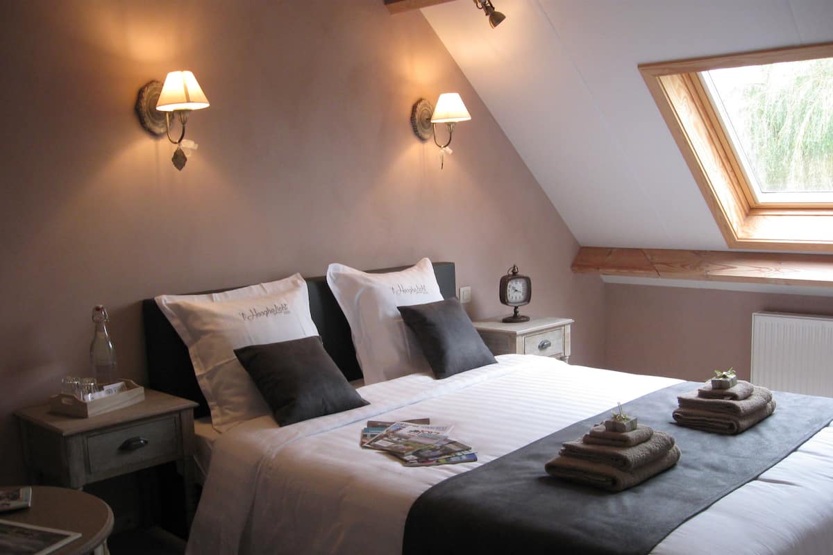 Une chambre à coucher comprenant un lit, deux tables de chevet, une commode, une horloge et une fenêtre donnant sur un paysage verdoyant.