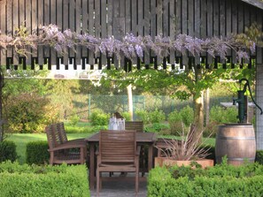 Een houten pergola met paarse bloemen, een houten tafel met stoelen en een waterpomp.
