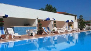 Une piscine avec des chaises longues et des parasols, un espace bar et un bâtiment avec un mur carrelé et des plantes en pot.