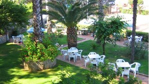 Une terrasse extérieure avec des chaises et des tables en plastique blanc, entourée d’une végétation luxuriante et de palmiers.