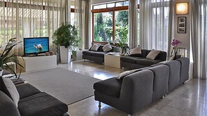 Un soggiorno moderno con un ampio divano grigio, una televisione a schermo piatto e una grande finestra con tende.