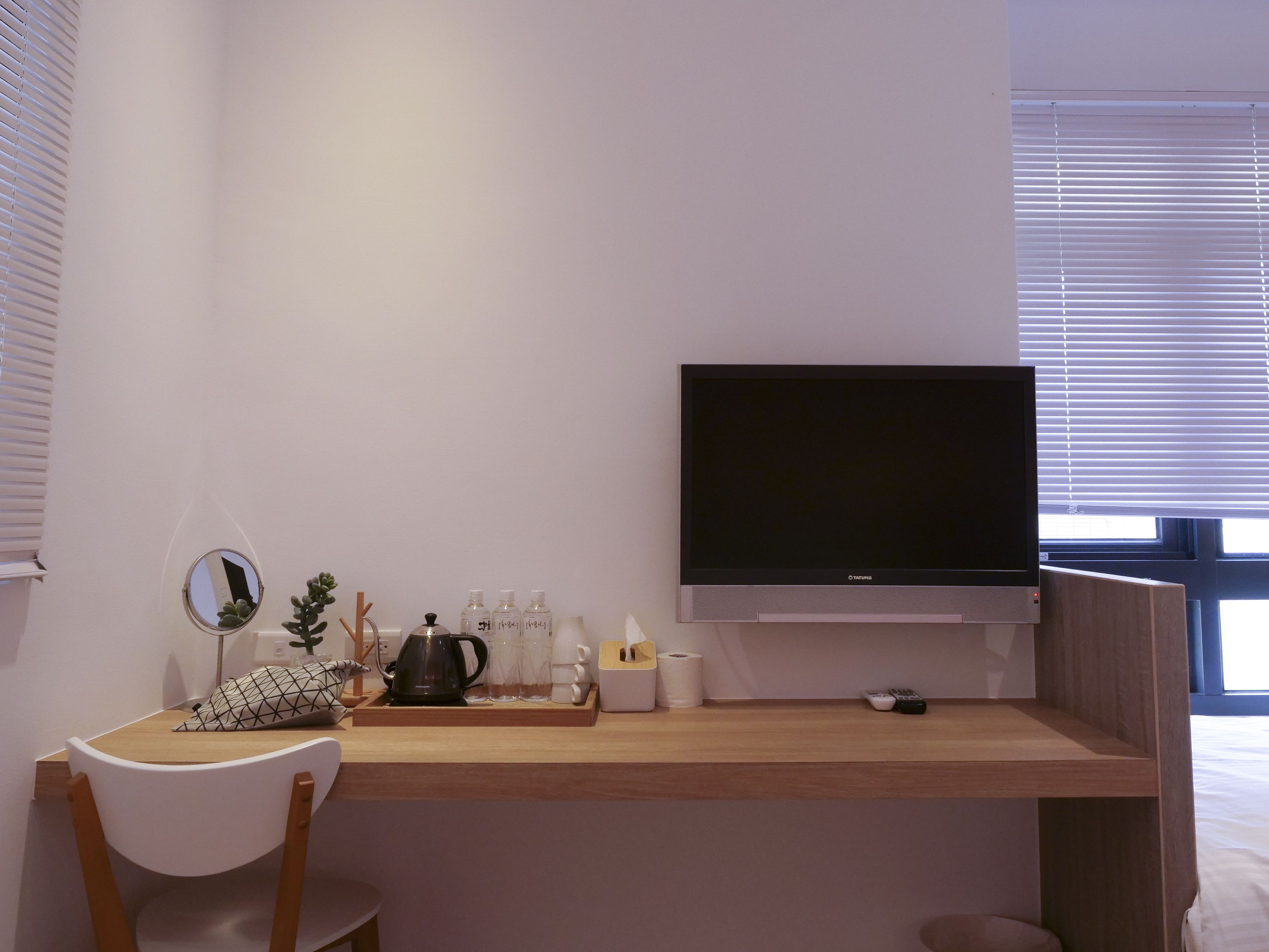 A modern room with a wooden desk, a flat-screen TV, a small mirror, and a window with blinds.