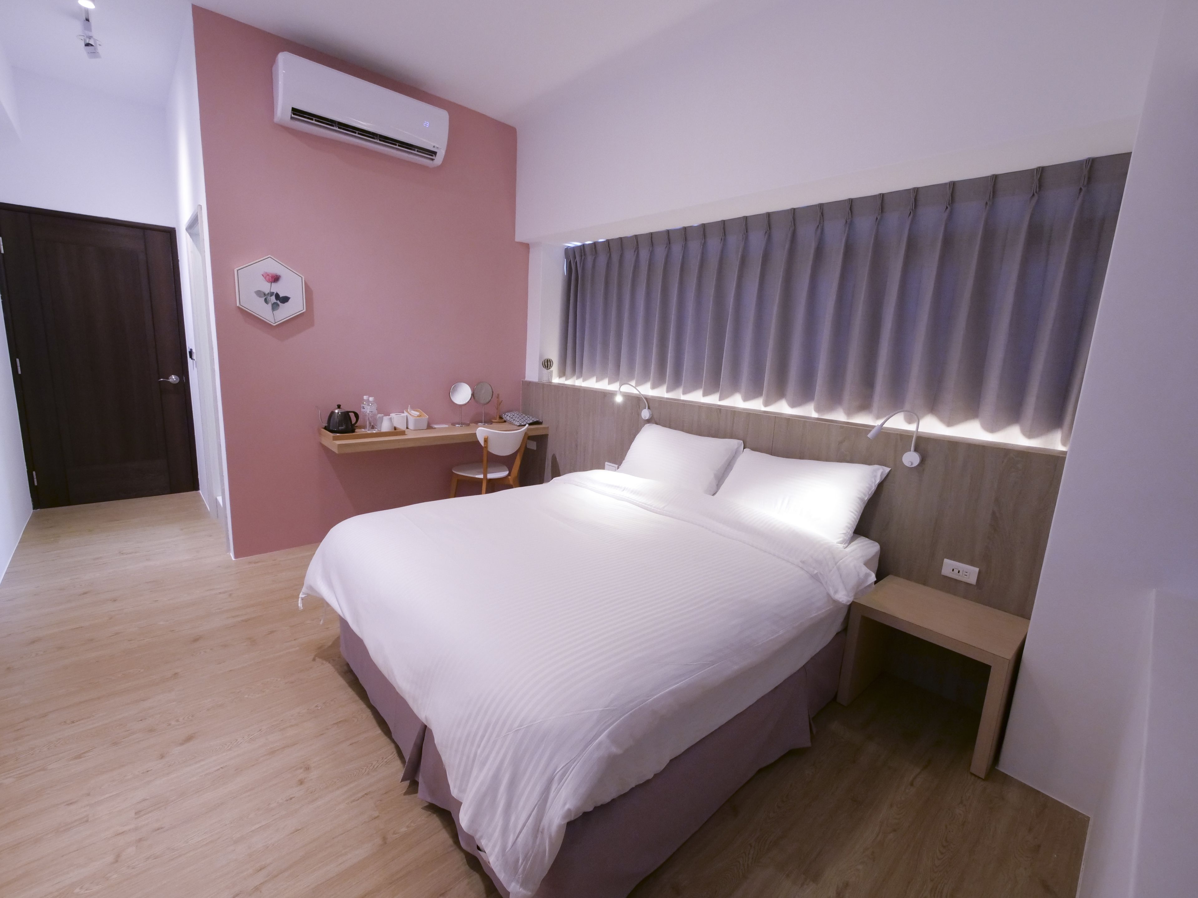 A modern hotel room with a large bed, bedside tables, a desk with a chair, an air conditioning unit, and a window with curtains.