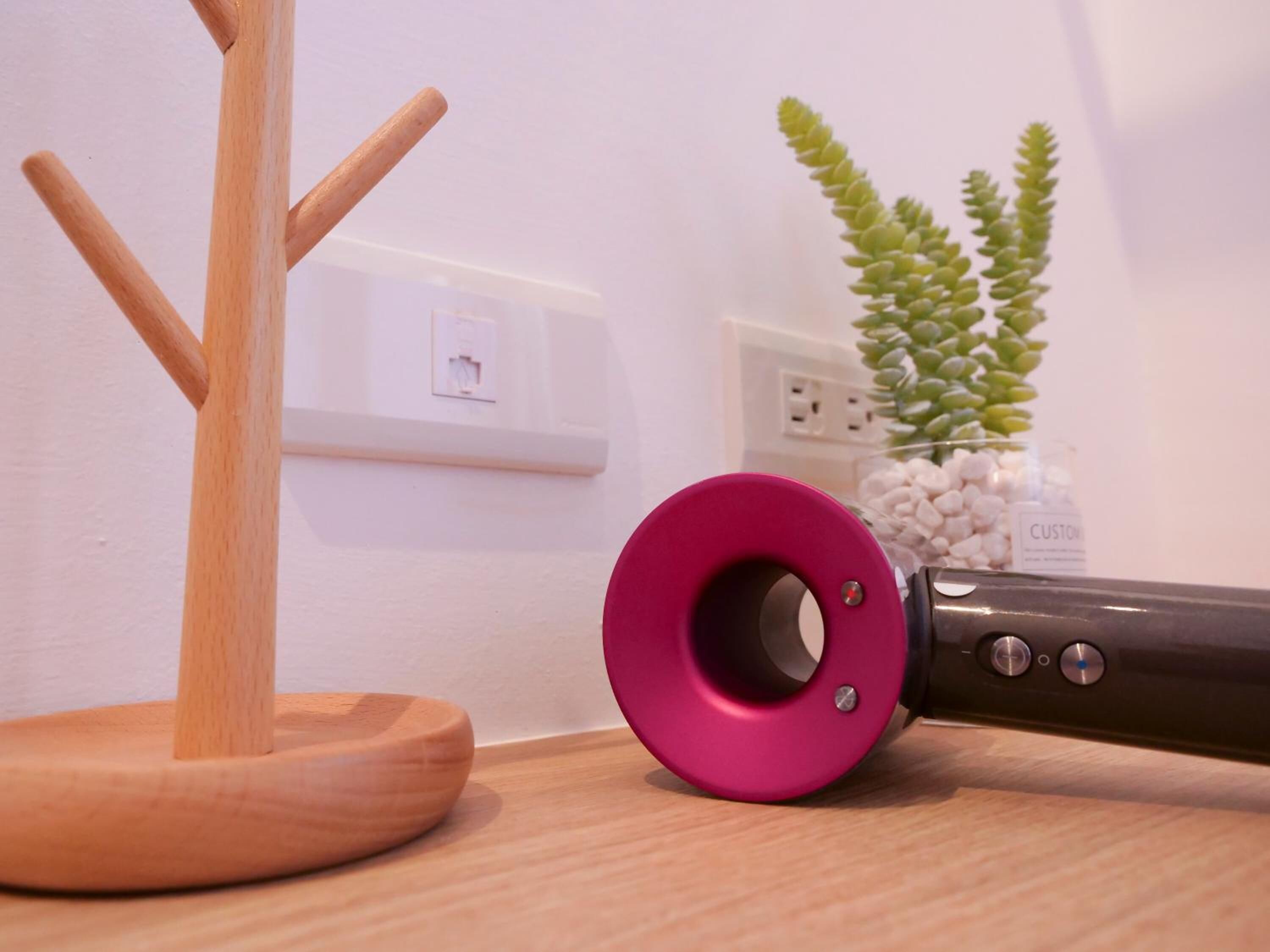 Un soporte de madera con un objeto rosa sobre una superficie de madera, un enchufe y una planta con guijarros blancos.