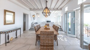 A spacious dining area with a long wooden table and wicker chairs, a hanging light fixture, and a glass door leading outside. - DreamLike Villas Mykonos (Mykonos)