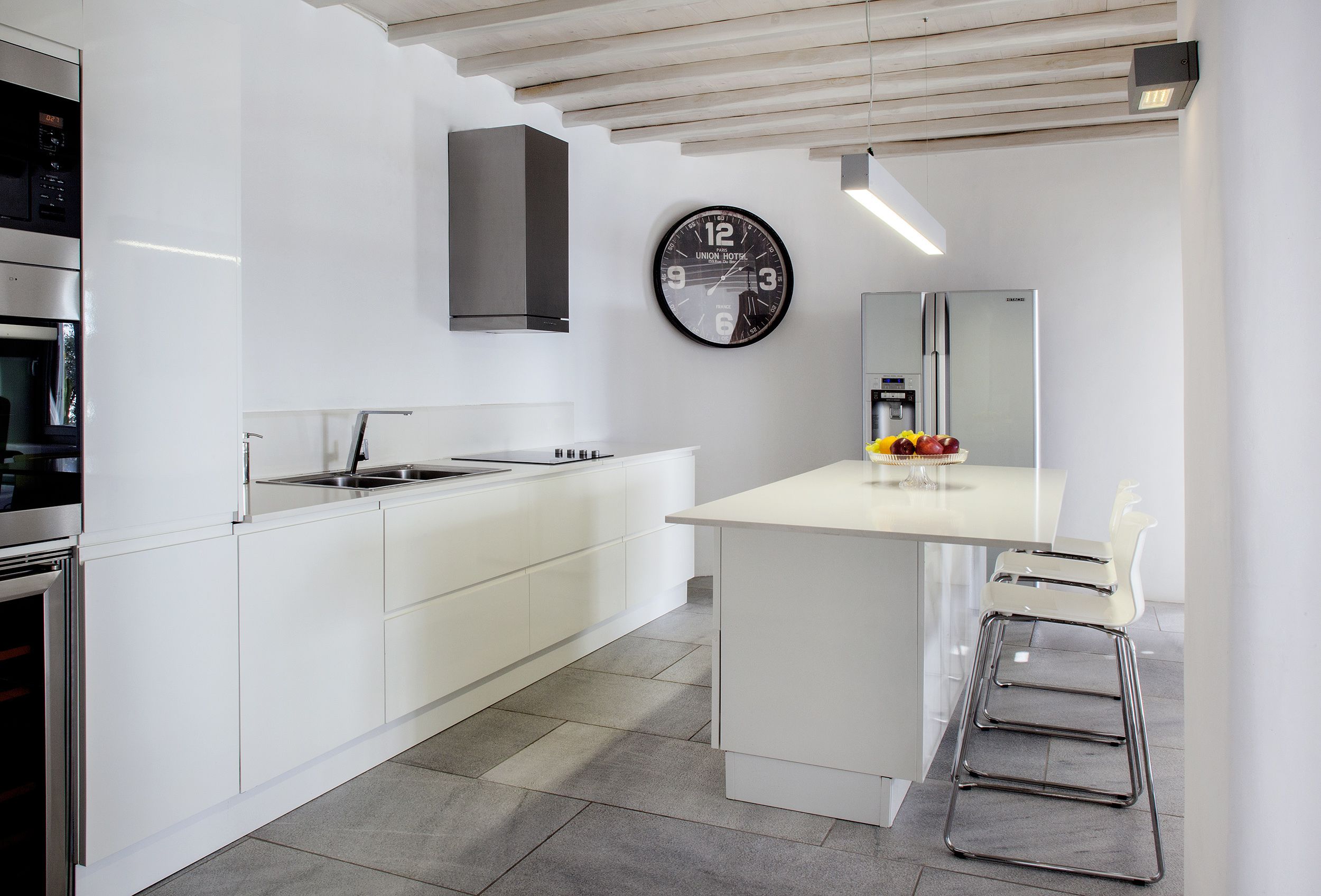 A modern kitchen with white cabinetry, stainless steel appliances, a central island with a sink, and a wall-mounted clock.