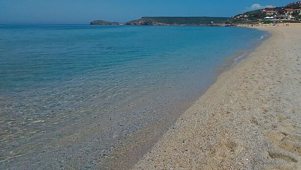 Ein Strand mit klarem Wasser und sandigem Ufer. Im Hintergrund sind Gebäude zu sehen.