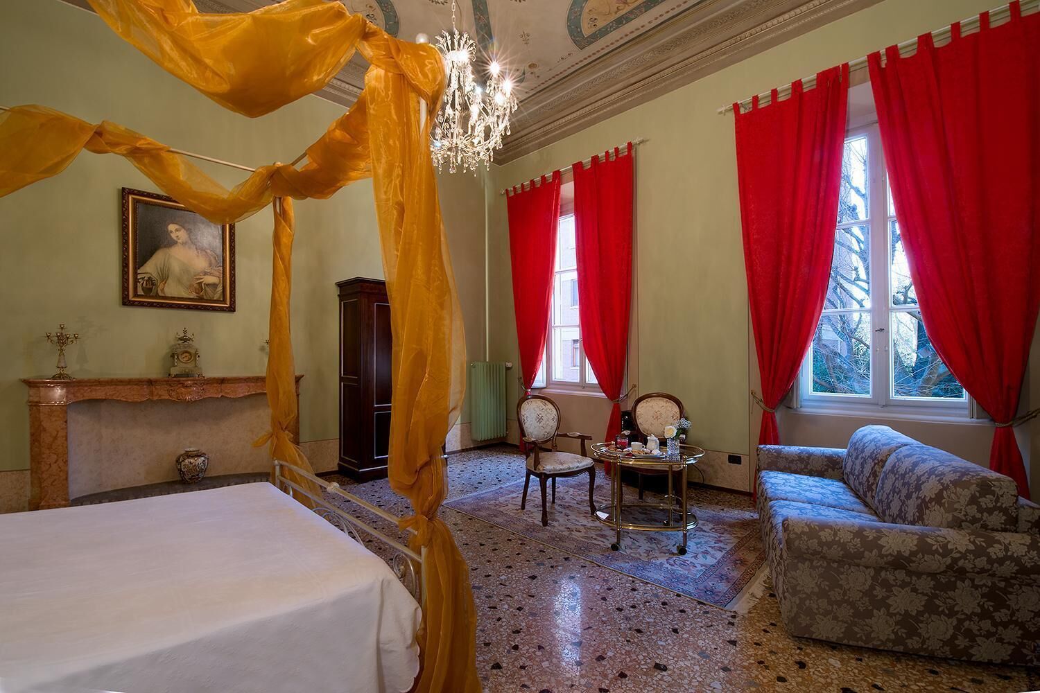 Een kamer met een hemelbed, een kroonluchter, rode gordijnen, een bank en een tafel met een dienblad.