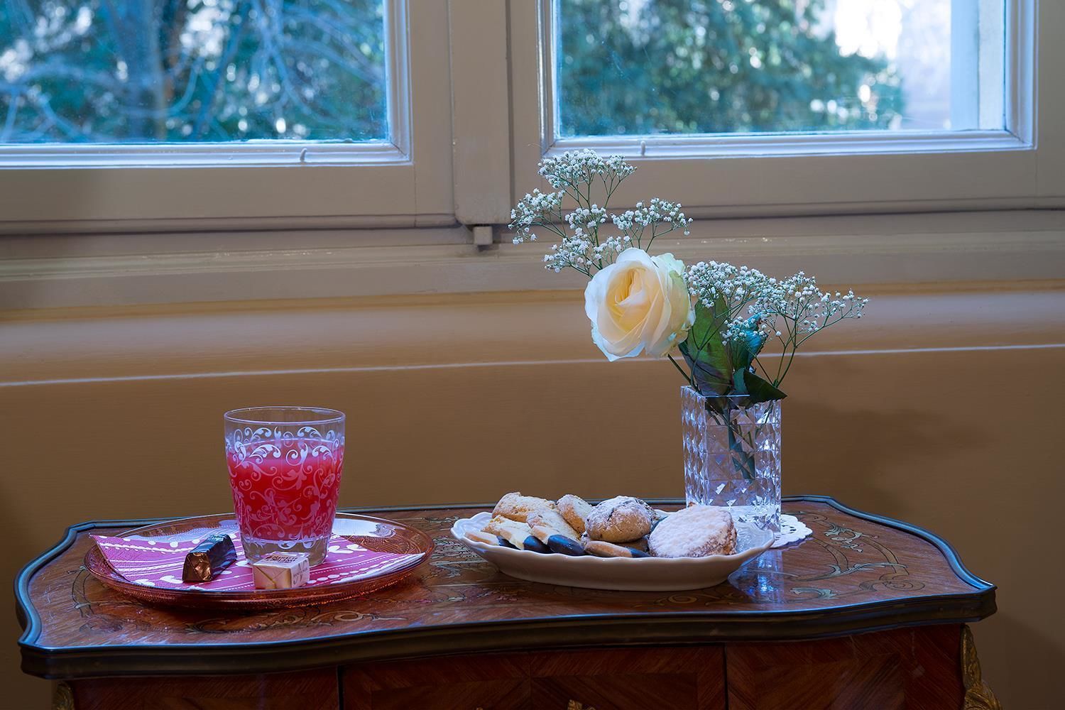 A table with a glass of pink drink, a plate of pastries, and a vase with flowers by a window.