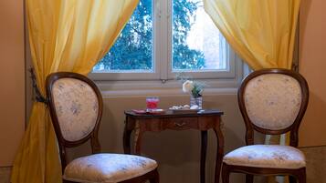 Two upholstered chairs with floral patterns, a small wooden table with a glass vase and a plate of cookies, and yellow curtains framing a window with a view of trees.