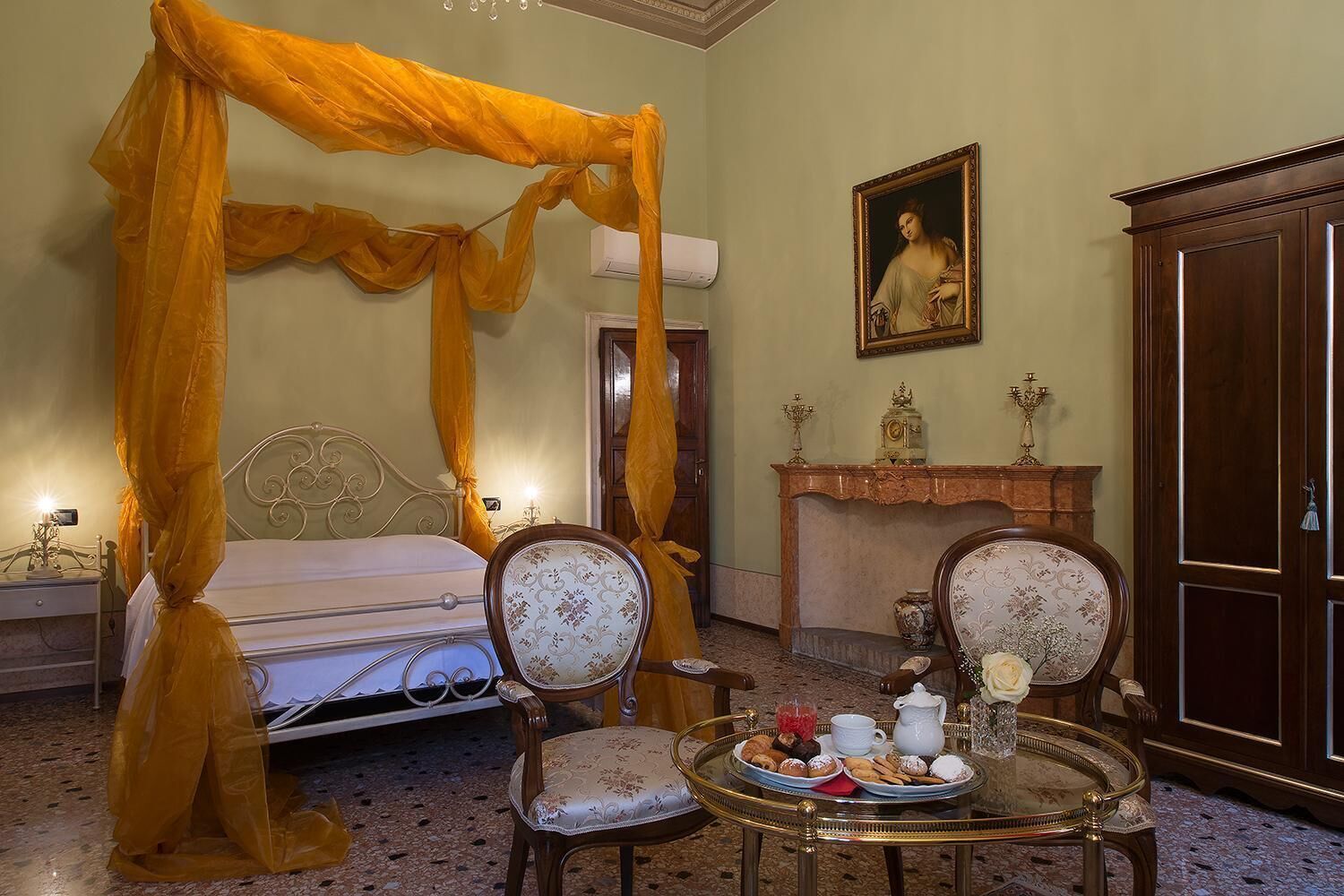 A room with a four-poster bed, two upholstered chairs, a round table with a tray of pastries, a fireplace, and a framed portrait on the wall.