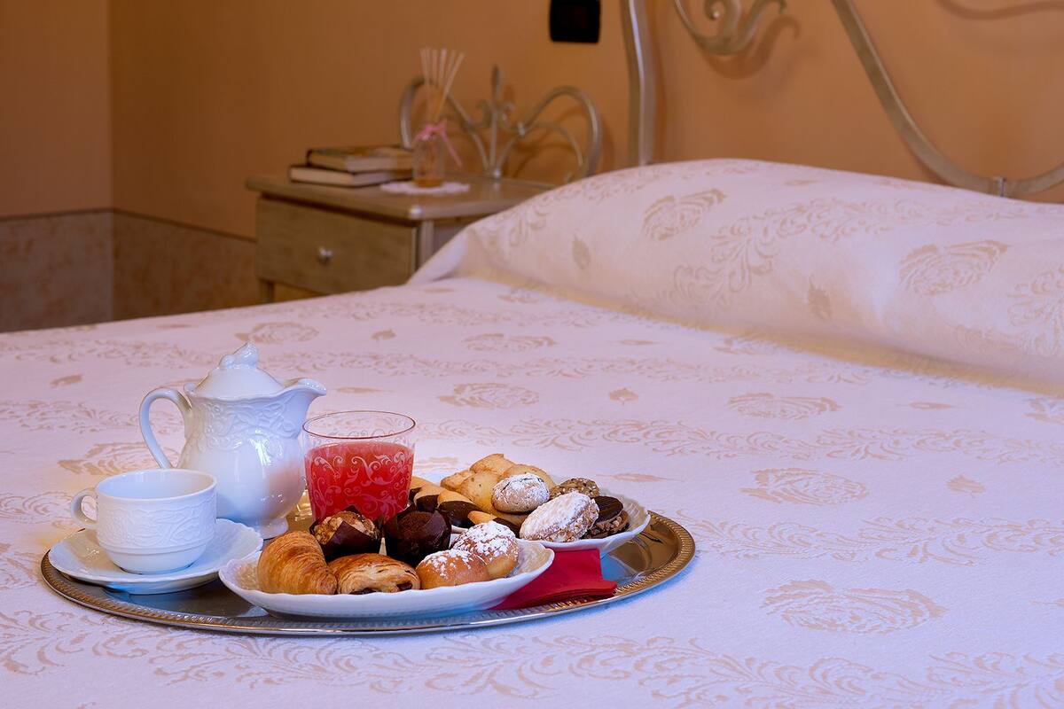 A neatly arranged breakfast tray on a bed with a white patterned bedspread, including croissants, pastries, a teapot, and a glass of red beverage.