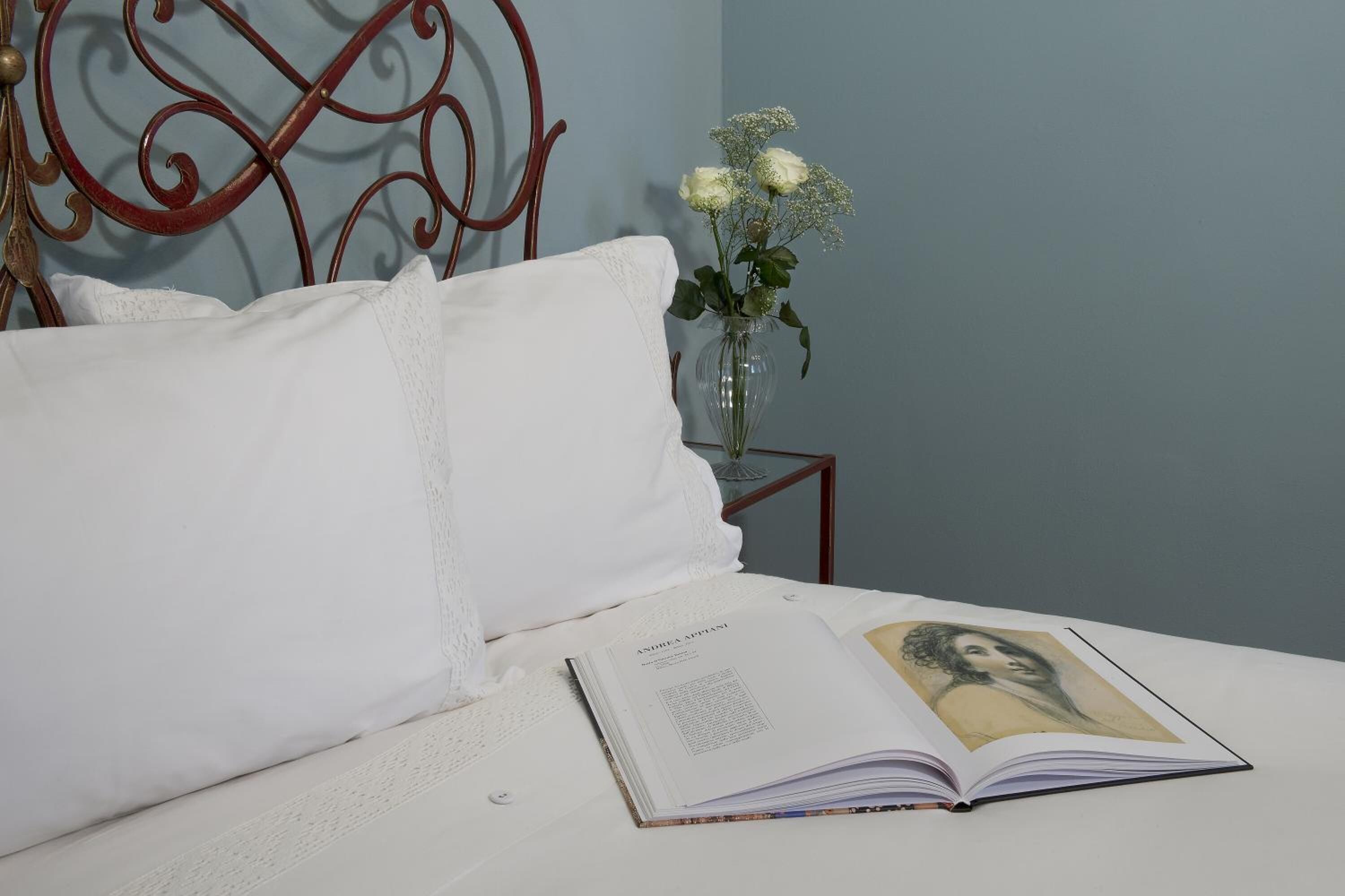 A neatly made bed with white linens, a book open on the bed, and a vase with flowers on a nightstand beside the bed.