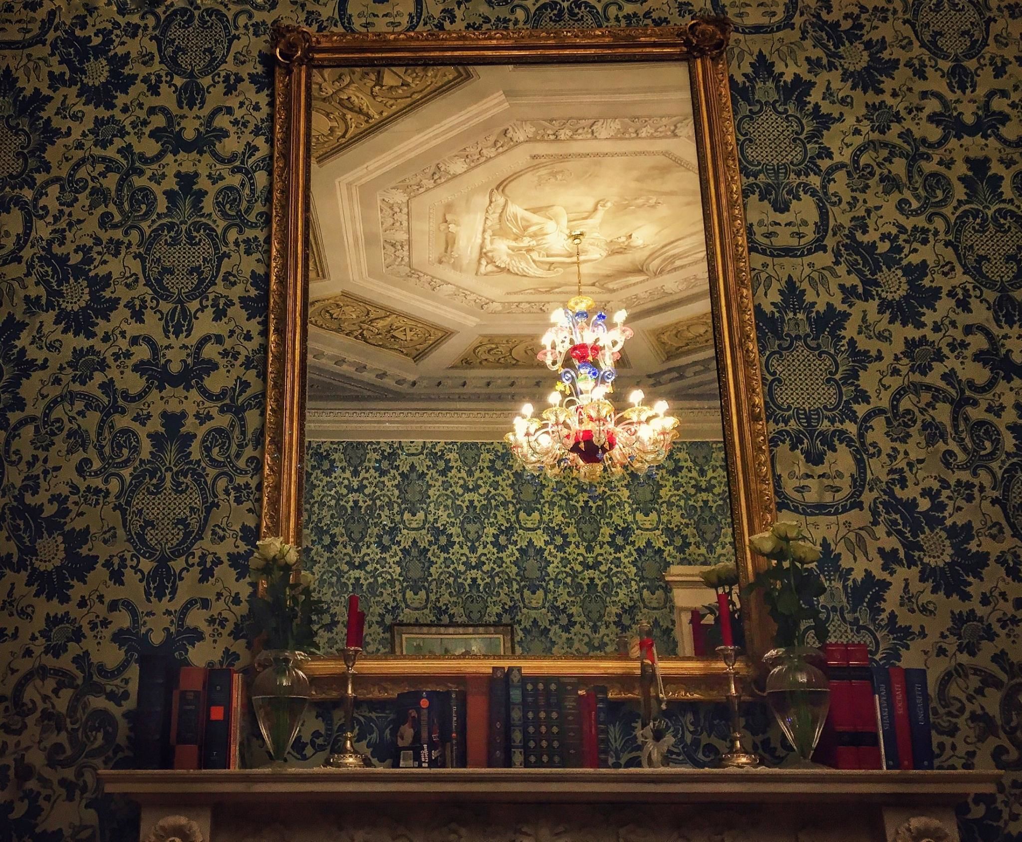 A room with a large ornate mirror, a chandelier, and patterned wallpaper.