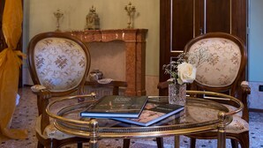 A room with two ornate chairs, a glass table with a book, a vase with flowers, and a fireplace with decorative items. - Salotto delle Arti (Modena)