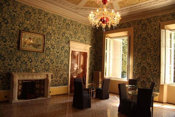 A room with patterned wallpaper, a fireplace, a chandelier, and a dining area with chairs and a table. - Salotto delle Arti (Modena)