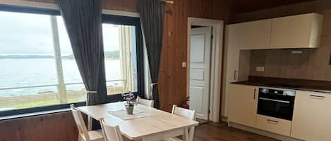 Una habitación amplia con suelo de madera, una mesa de comedor con sillas, una zona de cocina con armarios y electrodomésticos, y una ventana con cortinas.
