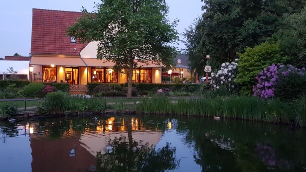 A hotel with a red-tiled roof, a garden with a pond, and a reflection of the building in the water. - Hotel Nierswalder Landhaus (Goch)