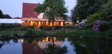 Um hotel com telhado de telhas vermelhas, um jardim com lago e o reflexo do edifício na água.