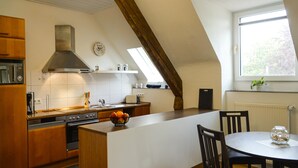 A modern kitchen with wooden cabinets, a central island, a built-in oven, and a large window providing natural light. - Hotel Nierswalder Landhaus (Goch)