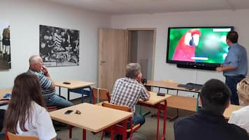Een klaslokaal met leerlingen zittend aan tafels, een leerkracht die voor de klas staat, en een groot scherm waarop een kleurrijk beeld te zien is.