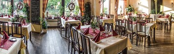 Restaurante com várias mesas preparadas para refeições, com piso de madeira e luz natural proveniente de grandes janelas.