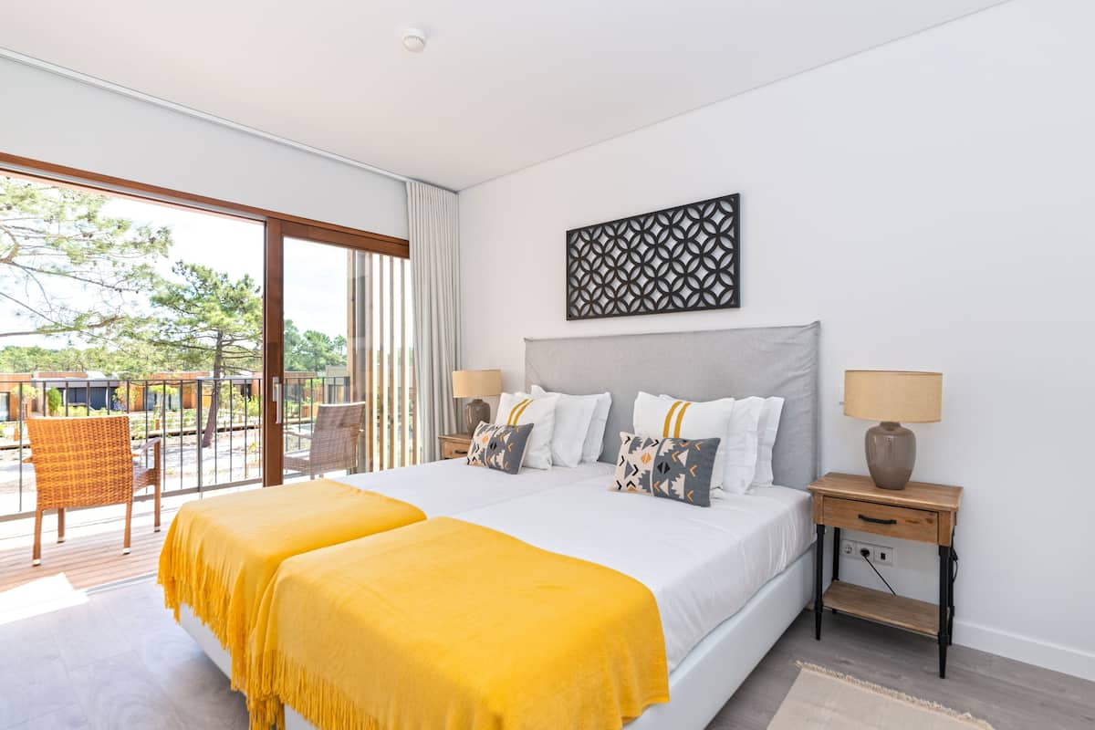 Un dormitorio con una cama grande, dos mesitas de noche, un adorno en la pared y un balcón con sillones de mimbre.