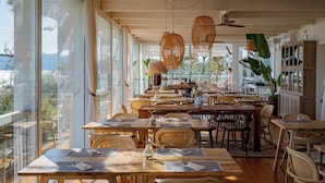 A spacious dining area with wooden tables and chairs, large windows offering views of the outdoors, and hanging rattan light fixtures. - Pestana Tróia (Grandola)