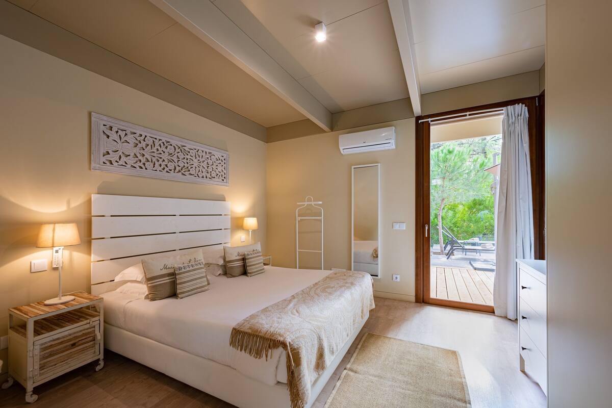 Un dormitorio con una cama grande, lámparas de noche, un adorno en la pared, un equipo de aire acondicionado y vistas al exterior a través de una puerta corrediza.