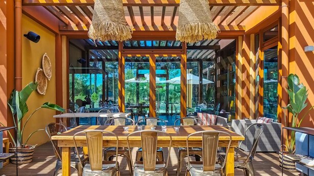 An outdoor dining area with a wooden table and metal chairs, surrounded by large windows offering a view of the outdoors. - Pestana Tróia (Grandola)