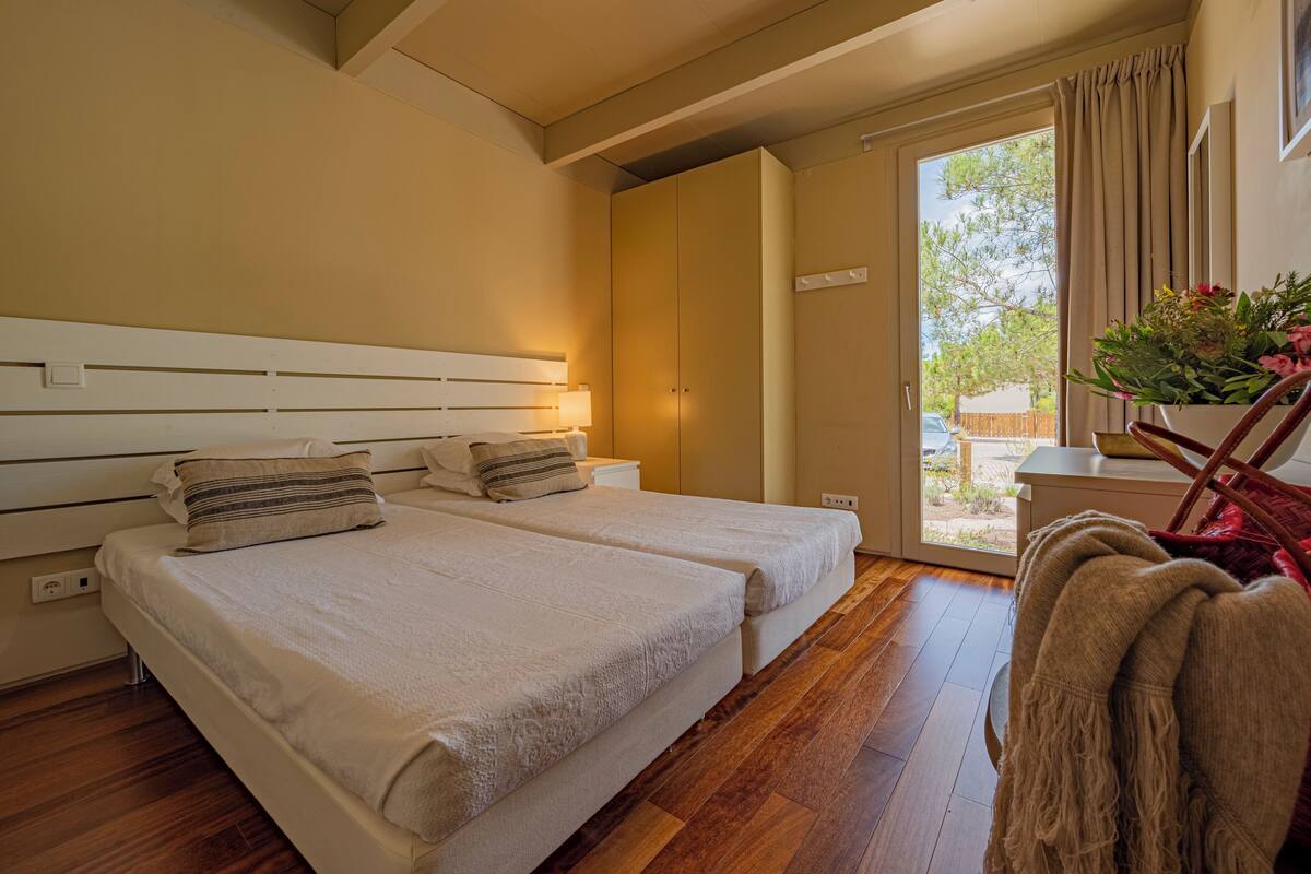 Un dormitorio con dos camas, suelo de madera, un ventanal con cortinas y una planta sobre una mesita.