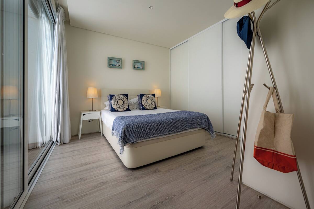 Un dormitorio con cama, lámparas de noche, una mesita de noche, una ventana con cortinas y un perchero en la pared.