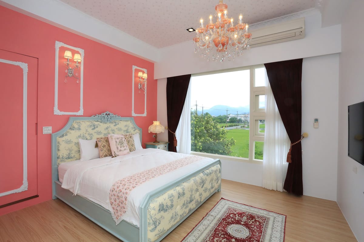 A bedroom with a bed, a chandelier, a window with curtains, and a view of greenery outside.
