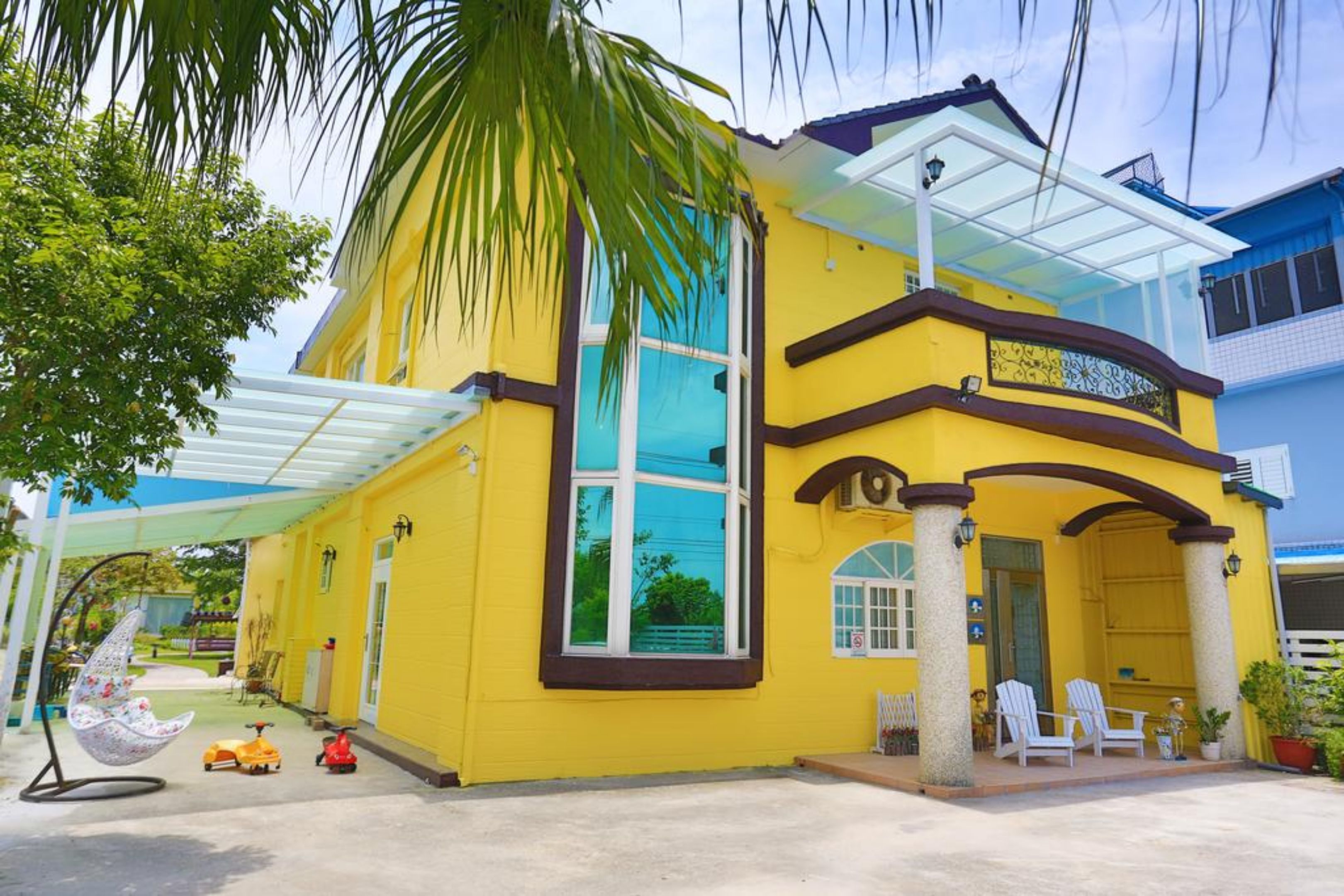 A yellow building with a large window, a covered porch, and a white chair outside.